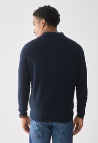 Marineblauwe gebreide trui met een geribbelde textuur, voorzien van een kraag en lange mouwen, gecombineerd met lichtblauwe spijkerbroek.