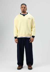 Gele oversized sweatshirt met een zwarte halslijn en een logo. Gecombineerd met donkerblauwe wijde broekspijpen en bruine veterlaarzen.