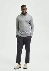 Maglione grigio con zip, pantaloni scuri e scarpe beige. Il maglione presenta un colletto alto e polsini a costine con una texture liscia.