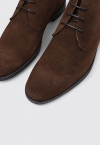 Botas de tobillo de ante marrón con textura suave, punta afilada, diseño con cordones y sutiles detalles de costura a lo largo de los bordes.