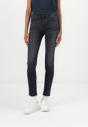 Person trägt dunkelgraue enganliegende Jeans, weiße Socken, weiße Turnschuhe und ein dunkelblaues Hemd und steht an einer schlichten weißen Wand.