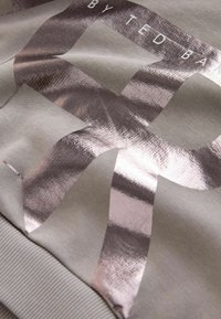 Lysegrå sweatshirt med et skinnende metalisk roseguld grafisk design og ribbet kant. Glat tekstur med markant brandingdetalje.
