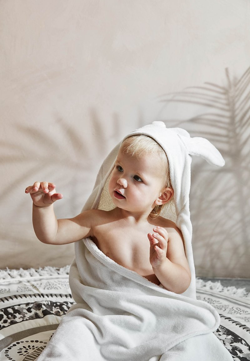 Valkoinen huppupyyhe, jossa on pupunkorvat, pehmeä tekstuuri, kietoutuu lyhythiuksisen lapsen ympärille, joka istuu kuvioidulla pinnalla, jossa on koristeellinen reuna.