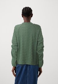 Pull vert texturé avec un motif à pois, manches longues et encolure ronde, porté avec une jupe en jean, vu de dos.
