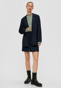 Navyblau gestreiftes Blazer- und Minirock-Set mit einem olivgrünen Oberteil, das Knöpfe, Taschen und schwarze klobige Stiefeletten hat.