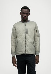 Chaqueta bomber acolchada en gris claro, con cuello redondo, cierre frontal con cremallera, bolsillos laterales y un bolsillo con cremallera en el brazo izquierdo. Textura suave.