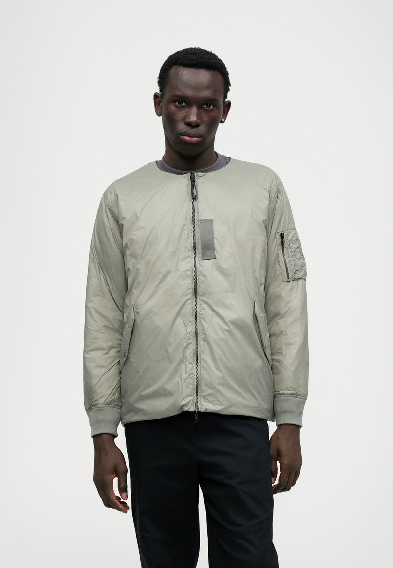 Chaqueta bomber acolchada en gris claro, con cuello redondo, cierre frontal con cremallera, bolsillos laterales y un bolsillo con cremallera en el brazo izquierdo. Textura suave.