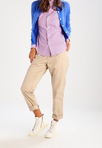 Modrý svetr, pruhovaná růžová a bílá košile, béžové nařasené kalhoty a bílé tenisky. Hladké textury a uvolněný střih po celém oblečení.