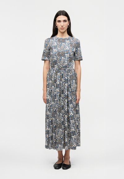 Femme aux longs cheveux foncés portant des boucles d'oreilles créoles et une robe midi à manches courtes bleue et blanche à fleurs, avec des ballerines noires, debout devant un fond blanc.