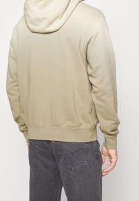 Felpa color khaki con schiena intera, polsini e orlo a costine. Presenta un cappuccio con coulisse. Abbinata a jeans scuri con tasche posteriori cucite.