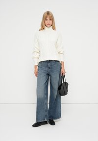 Maglione crema a trecce con collo alto, abbinato a jeans in denim blu a gamba larga. Borsa nera con angoli arrotondati e scarpe basse.