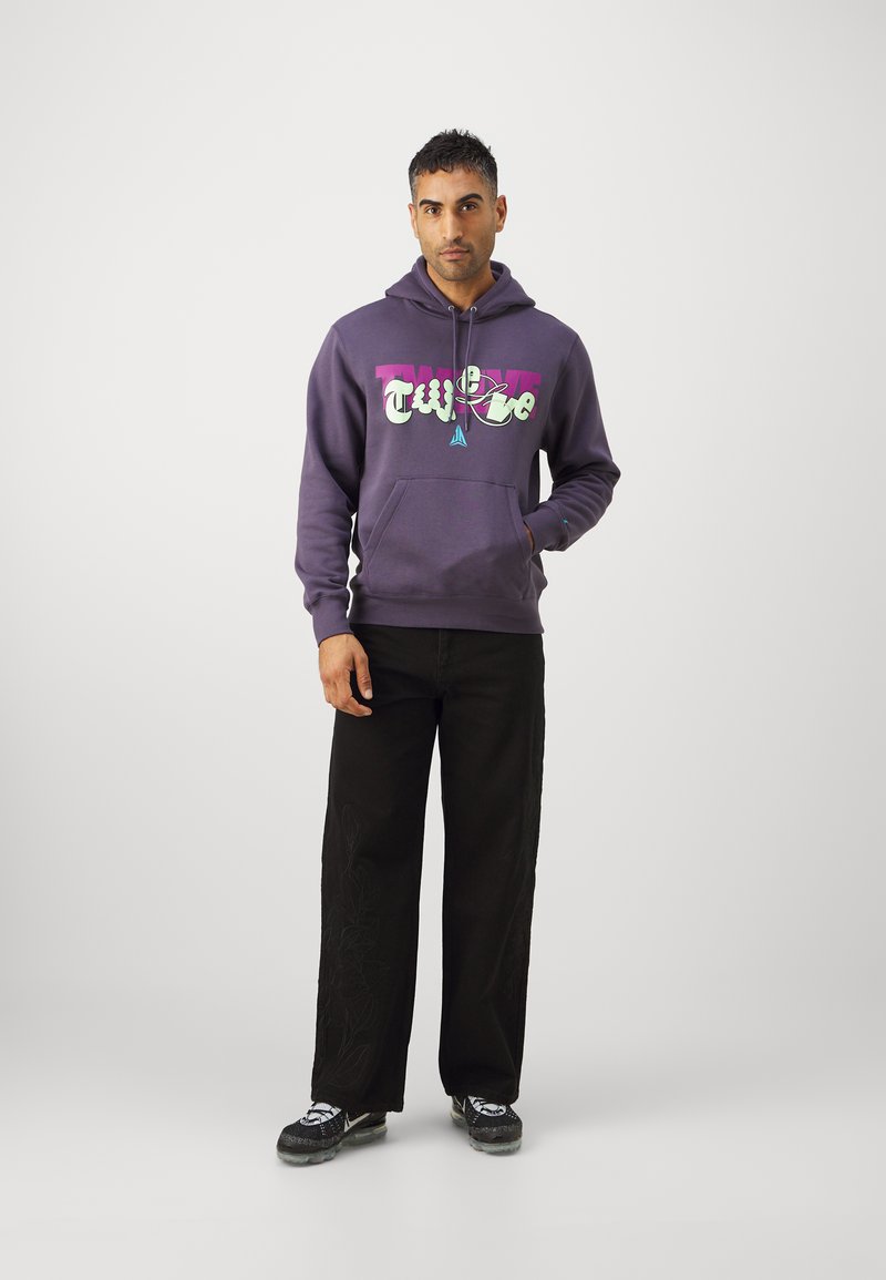 Mężczyzna stoi ubrany w fioletowy hoodie z grafiką, czarne szerokie spodnie z kwiatowym wzorem i czarne sneakersy, na jednolitym szarym tle.