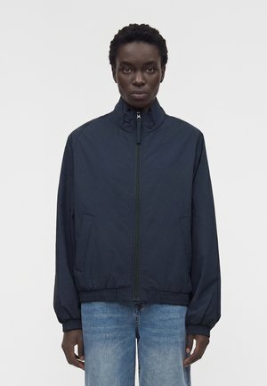 Personne portant une veste zippée bleu marine foncé avec col montant et un jean bleu clair sur un fond blanc uni.