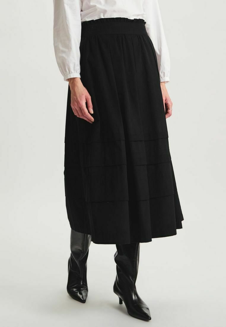 Jupe plissée noire en tissu texturé, taille élastique, arrivant au mollet, assortie à des bottes noires jusqu'aux genoux, créant un look superposé.