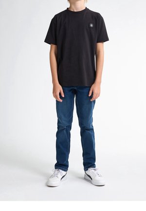 Camiseta negra de algodón con mangas cortas y cuello redondo. Presenta un pequeño parche con el logotipo en el pecho izquierdo. Combinada con vaqueros azules y zapatillas blancas.