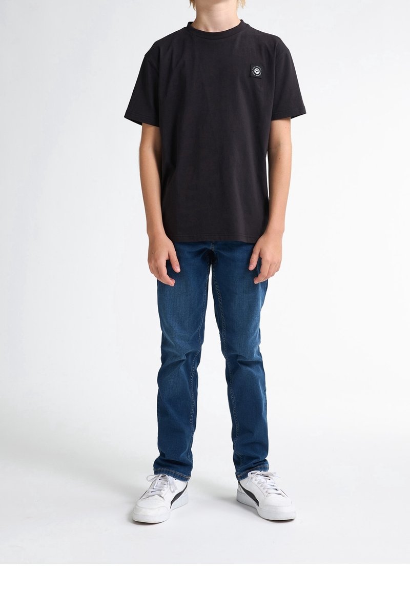 T-shirt noir en coton à manches courtes et col rond. Présente un petit patch logo sur le côté gauche de la poitrine. Associé à un jean bleu et des baskets blanches.