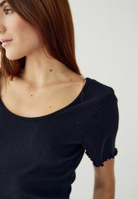 Melnā ribu adītā topo ar apaļu kakla izgriezumu, īsām pušķotām piedurknēm ar viļņotu maliņu un struktūru.