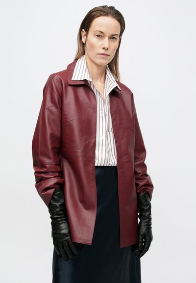 Γυναίκα με μπορντό δερμάτινο μπουφάν, λευκό πουκάμισο με κόκκινες ρίγες, μαύρη φούστα και μαύρα δερμάτινα γάντια, στέκεται μπροστά σε ένα απλό φόντο.