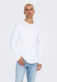 Bílá košile s dlouhým rukávem z bavlny, s kulatým výstřihem a mírně zaobleným lemem, v kombinaci s světle modrými džínami.