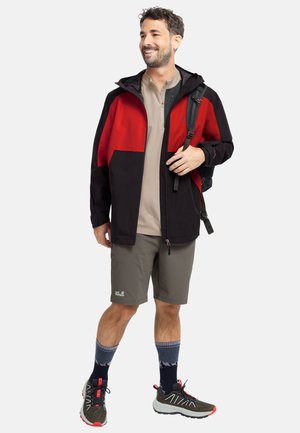Schwarze und rote wasserdichte Jacke mit Kapuze, beiges Hemd, graue Shorts, gemusterte Socken und dunkle Sportschuhe mit roten Akzenten.