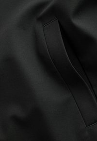 Črna tkanina z gladko teksturo, ki vključuje všit žep in subtilne detajle šivov. Celoten dizajn je minimalističen in elegantno zasnovan.