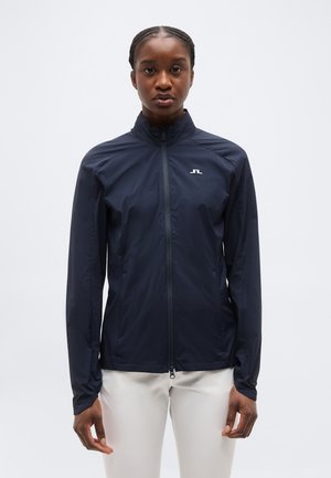 Γυναίκα που φοράει σκουρόχρωμο μπλε μπουφάν με φερμουάρ και μικρό λευκό λογότυπο στο στήθος και εκρού παντελόνι, στέκεται μπροστά από απλό φόντο.