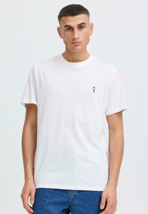 Young man with short hair wearing a plain white T-shirt with small embroidered figure on left chest and blue jeans, standing against white background.