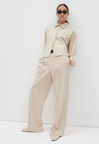 Beige, oversized shirt met een kraag en knoopsluiting, gecombineerd met losse broek. De look bevat zwarte accentdetails en platte schoenen.
