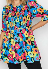 Nainen, joka on pukeutunut kirkkaaseen kukkamekkoon, jossa on sinisiä, vaaleanpunaisia, keltaisia ja oransseja kukkia mustalla taustalla. Hän poseeraa käsi lantiolla.