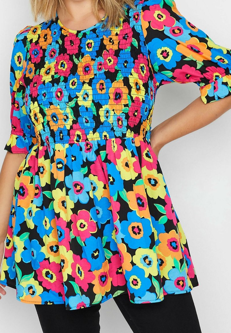 Nainen, joka on pukeutunut kirkkaaseen kukkamekkoon, jossa on sinisiä, vaaleanpunaisia, keltaisia ja oransseja kukkia mustalla taustalla. Hän poseeraa käsi lantiolla.