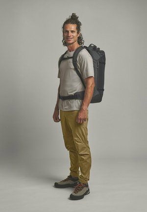 Man wearing a gray t-shirt, tan pants, hiking shoes, and carrying a large black hiking backpack, standing against a plain background.