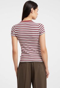 Donna di spalle che indossa una camicia a maniche corte bianca, rossa e nera a righe orizzontali con pantaloni marroni.