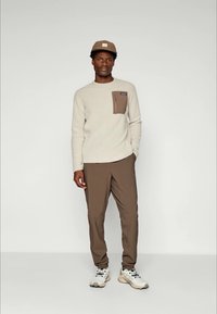 Pull en maille texturée beige avec une poche carrée marron ; pantalon slim marron ; baskets claires ; casquette marron avec un détail de logo.