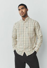 Camisa de cuadros crema con líneas verdes y naranja, con un bolsillo en el pecho y un logotipo en el lado izquierdo. Combinada con pantalones oscuros.