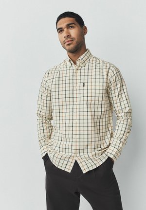 Camisa de cuadros crema con líneas verdes y naranja, con un bolsillo en el pecho y un logotipo en el lado izquierdo. Combinada con pantalones oscuros.