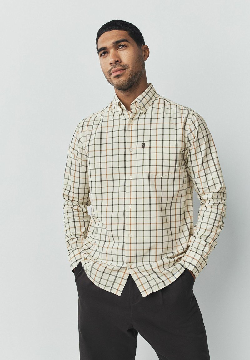 Camisa de cuadros crema con líneas verdes y naranja, con un bolsillo en el pecho y un logotipo en el lado izquierdo. Combinada con pantalones oscuros.