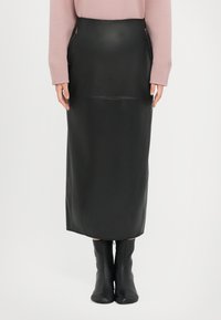 Fustă midi din piele neagră, cu fermoare laterale, textură netedă și siluetă dreaptă. Poartă cizme negre ajustate până la genunchi.