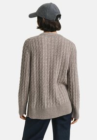 Taupe gebreid sweater met een gestructureerd kabelpatroon, ribbels aan de onderkant en lange mouwen, gecombineerd met donkere denim broeken. Achteraanzicht getoond.