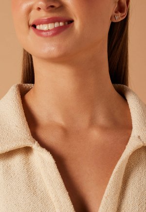 Lächelnde Frau mit herzförmigen Ohrsteckern und einem strukturierten cremefarbenen Oberteil mit Kragen auf beigem Hintergrund.