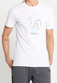 Mies, który ma na sobie białą koszulkę z stylizowanym białym i szarym kogutem oraz literami "FFF" na klatce piersiowej, w zestawieniu z ciemnoszarymi spodniami.