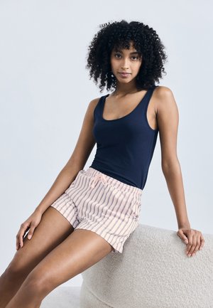 Femeie cu păr creț așezată, purtând un maiou bleumarin și pantaloni scurți roz deschis cu dungi, pe un fundal simplu, deschis la culoare.