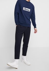 Muž v námořnickém modrém svetru ellesse, tmavých zkrácených kalhotách, bílých ponožkách a bílých teniskách, stojící s rukama u kapsy.