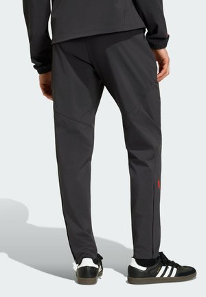 Uomo con pantaloni sportivi neri e scarpe da ginnastica Adidas nere con strisce bianche, visto di spalle, in piedi su uno sfondo chiaro.