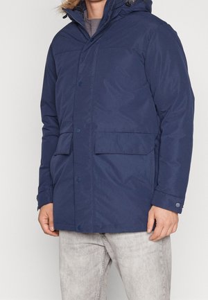 Man draagt een marineblauwe winterjas met capuchon en klepzakken, gecombineerd met lichtgrijze jeans, staand tegen een effen achtergrond.