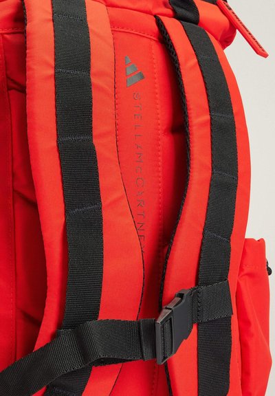 Red backpack with black straps, padded texture, and adjustable buckles. Features branding: "STELLA McCARTNEY" on the side.