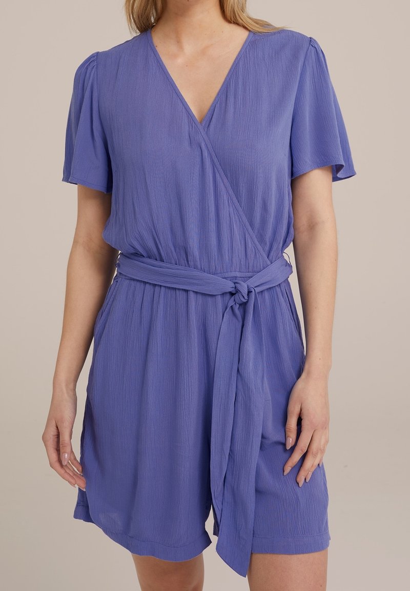 Femme portant une robe portefeuille lavande à manches courtes et col en V, nouée à la taille avec une ceinture lâche, se tenant devant un fond neutre.