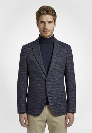 Mann mit hellbraunen Haaren und Bart, der einen strukturierten dunkelblauen Blazer über einem marineblauen Rollkragenpullover und beigen Hosen trägt, steht vor einem schlichten Hintergrund.