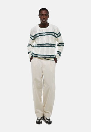 Pull en maille torsadée crème avec des rayures vertes, associé à un pantalon plissé crème et des baskets camouflées noires. Le mannequin se tient avec les mains dans les poches.