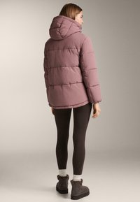 Kurtka puchowa w delikatnym kolorze mauve, z kapturem, pikowanym wzorem i nieco dłuższym tyłem. Połączona z ciemnymi legginsami i butami ocieplanymi futerkiem.