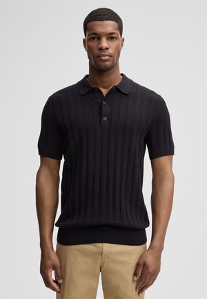 Man draagt een zwart, geribd, poloshirt met korte mouwen en beige broek, staand tegen een effen lichtgrijze achtergrond.